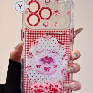 Pode incluir: Capa de telefone transparente com um padrão xadrez vermelho e branco e um design de "Strawberry Cake". A capa apresenta glitter e pequenos enfeites em forma de bolo. O design inclui um bolo rosa com laços e corações.