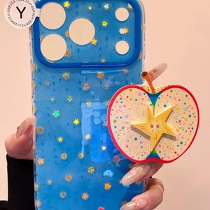 Peut inclure: Coque de téléphone bleue avec un motif d'étoiles et une poignée en forme de pomme. L'étui a une bordure transparente et un intérieur bleu avec des étoiles dorées et irisées. La poignée de la pomme est blanche, rouge et jaune.