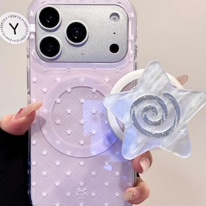 May include: A clear phone case with a lavender background and a pattern of small white stars. A white and silver star-shaped phone grip is attached. The phone case has a clear border.