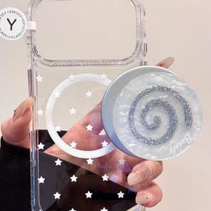 May include: Clear phone case with a white star pattern and a silver and white pop socket. The pop socket has a glitter swirl design and the word "Beauty" printed on it.