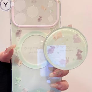 May include: A clear phone case with a light green and pink border, decorated with illustrations of bunnies and clovers. A matching pop socket with the same design is attached. The case has a circular cutout.
