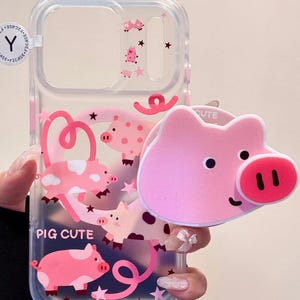 Puede incluir: Funda de teléfono transparente con ilustraciones de cerdos rosas y el texto "PIG CUTE". Se adjunta un agarre para teléfono con forma de cerdo rosa. La funda presenta estrellas rosas y diseños de cerdos.