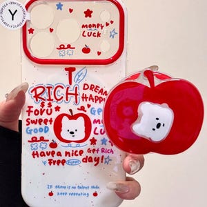 Könnte beinhalten: Eine weiße Handyhülle mit rotem Rand und einem roten, apfelförmigen Handyhalter. Die Hülle zeigt die Wörter "RICH", "DREAM", "LUCK" und "FREE" mit niedlichen Illustrationen. Der Apfelgriff hat ein weißes Bären-Design.