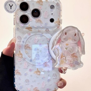 Puede incluir: Funda de teléfono transparente con un patrón de conejos y flores. La funda tiene un soporte transparente con un diseño de conejito. La parte trasera de la funda es transparente con un patrón de conejos y flores.