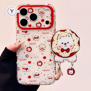 Könnte beinhalten: Transparente Handyhülle mit rotem Rand, verziert mit Cartoon-Bären, Äpfeln und dem Schriftzug "happy puppy". Ein Anhänger mit Bär- und Apfeldesign ist angebracht.