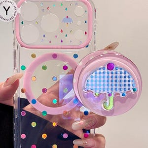 May include: Clear phone case with a pink border and colorful polka dots. The case features a pink pop socket with an umbrella design. The case also has a pink ring around the camera area and a small umbrella design.