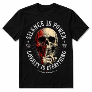 K&ouml;nnte beinhalten: Schwarzes T-Shirt mit einem Totenkopf-Design. Der Sch&auml;del ist geteilt, eine H&auml;lfte rot und die andere beige. Der Text um den Sch&auml;del lautet "SILENCE IS POWER" und "LOYALTY IS EVERYTHING". Zus&auml;tzlicher Text enth&auml;lt "SPEAK LESS", "TRUST FEW" und "NO NOISE. NO WEAKNESS."