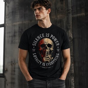 K&ouml;nnte beinhalten: Schwarzes Rundhals-T-Shirt mit einem Totenkopf-Motiv und dem Text "SILENCE IS POWER" in einem kreisf&ouml;rmigen Design. Der Sch&auml;del hat rote und beige Farbt&ouml;ne, mit dem Text "SPEAK LESS" und "TRUST FEW".