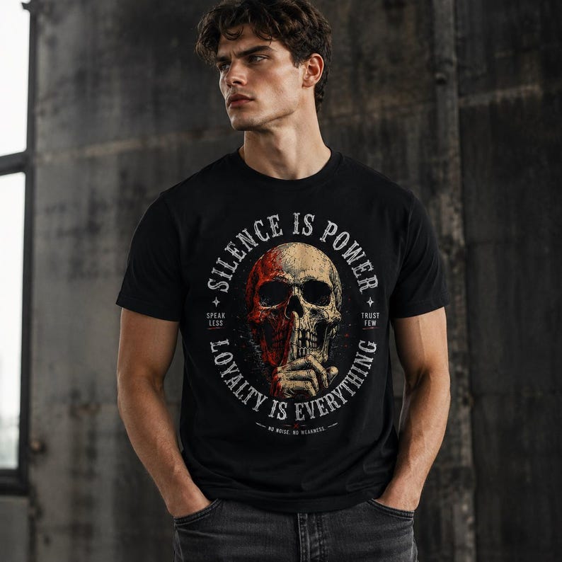 K&ouml;nnte beinhalten: Schwarzes Rundhals-T-Shirt mit einem Totenkopf-Motiv und dem Text "SILENCE IS POWER" in einem kreisf&ouml;rmigen Design. Der Sch&auml;del hat rote und beige Farbt&ouml;ne, mit dem Text "SPEAK LESS" und "TRUST FEW".