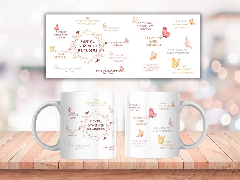 May include: White ceramic coffee mug with the text "Mental Strength Reminders" and inspirational phrases. The design includes butterflies and floral elements in shades of pink and gold. The mug is on a wooden surface.