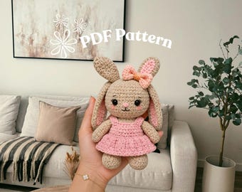 May include: A beige crochet bunny doll with long ears, wearing a pink dress and a small peach-colored bow on its head. The bunny has embroidered black eyes, a pink nose, and rosy cheeks. The text PDF Pattern is visible in the upper portion.