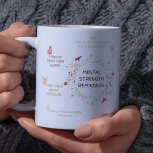 May include: White ceramic mug with the text "Mental Strength Reminders" surrounded by floral and butterfly illustrations. Additional phrases include "I choose peace over worry" and "Stay calm under pressure."