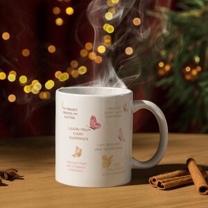 May include: A white ceramic mug with a curved handle, featuring inspirational text and butterfly illustrations. The mug is filled with a steaming beverage, and cinnamon sticks are placed on a wooden surface.