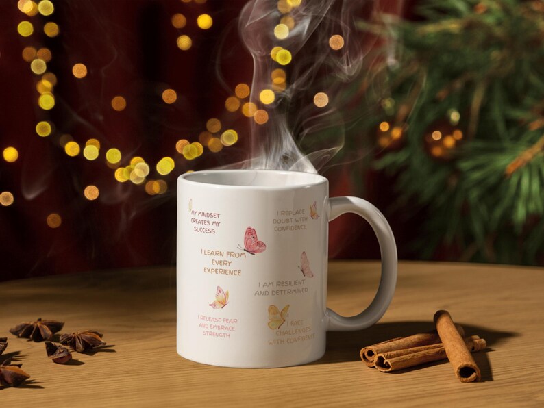 May include: A white ceramic mug with a curved handle, featuring inspirational text and butterfly illustrations. The mug is filled with a steaming beverage, and cinnamon sticks are placed on a wooden surface.
