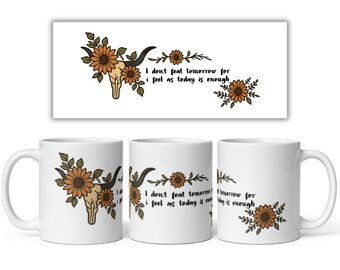 May include: A white ceramic mug featuring a graphic of a cow skull adorned with brown sunflowers and green leaves. The text reads, I don't feat tomorrow for i feel as today is enough, written in a black script font next to the floral skull design.