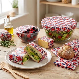 May include: A collection of reusable food wraps featuring a red, white, and blue Union Jack pattern. The wraps cover glass bowls filled with berries and salad, a block of cheese, a sandwich, and a loaf of bread on a wooden surface.