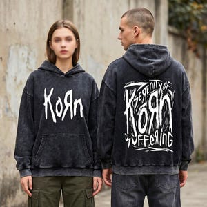 May include: Two people wearing black hoodies with the band name "KoЯn" in white, stylized lettering. The back of one hoodie features the text "Serenity of Suffering" in a similar font. The hoodies have a washed-out, vintage look.
