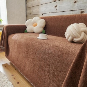 Puede incluir: Un sofá marrón con una manta a juego. Los cojines decorativos incluyen una almohada blanca en forma de flor con un centro amarillo y una almohada de nudo color crema. Una taza y un platillo blancos están sobre el sofá. El fondo es una pared de madera.