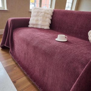 Könnte beinhalten: Ein weinrotes Sofa mit passendem Überwurf und einem weißen, flauschigen Kissen. Eine weiße Tasse mit Untertasse steht auf dem Sofa. Der Raum ist gut beleuchtet.