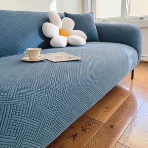 May include: A blue sofa with a geometric pattern. A white flower-shaped cushion with a yellow centre sits on the sofa, along with a small cushion and a teacup on a saucer. A magazine is also on the sofa.