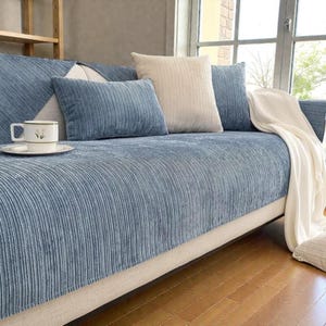 Pode incluir: Uma capa de sofá de veludo cotelê azul com almofadas a condizer em azul e bege. Uma manta branca está sobre o braço. Uma chávena e pires estão no sofá. O sofá está em frente a uma janela.