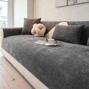 Puede incluir: Una funda de sofá gris con cojines a juego y un cojín decorativo en forma de nudo. Una taza y un platillo blancos están sobre la funda, junto con una pequeña tarjeta. El sofá tiene una base color crema.