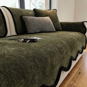 May include: A green sofa with matching pillows and a decorative cover. The sofa has a textured, ribbed fabric with black trim. A small gray pillow and a black purse with a silver chain are on the sofa.