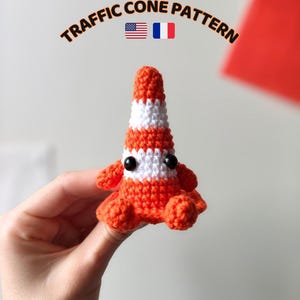 Peut inclure: Une petite figurine de cône de signalisation crochetée avec des rayures horizontales orange et blanches, dotée de deux yeux de sécurité en plastique noir et de petits bras et jambes crochetés orange. Le texte TRAFFIC CONE PATTERN est imprimé au-dessus.