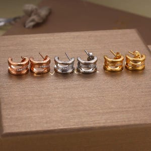 May include: Six pairs of hoop earrings in rose gold, silver, and gold. Each earring has a layered design with small, clear gemstones. The earrings are displayed on a brown surface, showcasing their different metallic finishes.