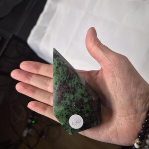 May include: A hand holds a faceted gemstone with a green and maroon pattern. The stone is triangular, with a flat base and a pointed top. A small white sticker with the number "40" is affixed to the stone.