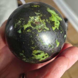 May include: A dark, spherical object with a polished surface. The sphere has a mottled pattern of black, dark green, and bright green hues. The object is held in a hand, with a silver ring visible on a finger.