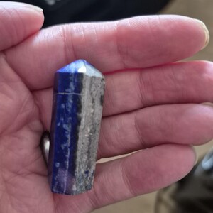 May include: A hexagonal crystal point made of lapis lazuli, featuring a deep blue colour on one side and a contrasting grey, textured mineral inclusion on the other. The stone has a polished finish and a pointed top, displaying natural variations.