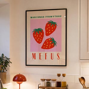 May include: A framed wall art print featuring three red strawberries with green leaves on a light pink background. The text "MARCHNAD FFRWYTHAU" appears in green at the top, and the word "MEFUS" is printed in large red capital letters at the bottom.