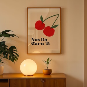 May include: A rectangular framed wall art print featuring two red cherries with green leaves on a light beige background. Below the cherries, the black text reads Nos Da Caru Ti. The print is displayed in a thin wooden frame on a plain wall.