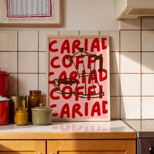 May include: A framed rectangular wall art print featuring a light pink background with the repeating red text "CARIAD COFFI" and a dark brown line drawing of a moka pot and a coffee cup centred on the print, encased in a light wood frame.