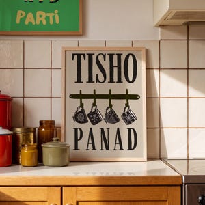 May include: A rectangular framed wall art print featuring a light beige background with the black text TISHO at the top and PANAD at the bottom. Between the words is a dark green horizontal rack with four hooks, each holding a black tea cup.