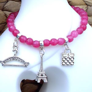 Pode incluir: Uma pulseira de contas redondas e facetadas em rosa choque com três berloques de metal prateado. Os berloques incluem um cabide decorativo, uma miniatura da Torre Eiffel e uma bolsa quadrada com o número 5 gravado na parte frontal.