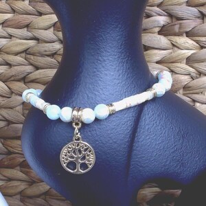 May include: A beaded bracelet featuring light blue and white stone beads interspersed with small gold-toned metal spacers. A circular gold-toned metal charm depicting a tree of life design hangs from the center of the bracelet, which is displayed on a dark blue stand.