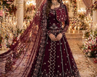 May include: A burgundy formal gown with intricate gold embroidery and a matching bolero jacket. The gown features a long, flowing skirt and a sheer veil. The background includes floral arrangements and decorative lighting, creating an elegant atmosphere.