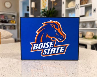 May include: A rectangular sign with a blue background featuring the Boise State logo, which consists of a stylized orange and blue horse head silhouette above the white text Boise State with an orange outline on a blue field.