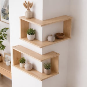 May include: Two light-colored wooden corner shelves mounted on a white wall. The top shelf holds a small vase with dried grasses, a potted green plant, a round stone-like object, and a wooden bowl. The bottom shelf holds a potted plant and a round object.