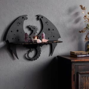 May include: A black wall-mounted shelf shaped like a dragon with outstretched wings. The wings feature cut-out star patterns. A flat horizontal surface holds a lit white tea light candle, two tall purple crystal points, and two smaller light-colored stones.
