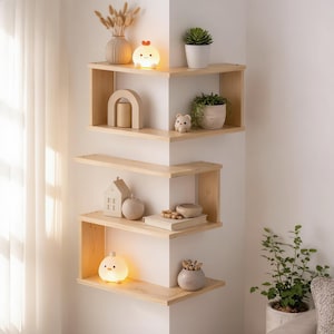 May include: A light-colored wooden corner shelving unit mounted on a wall, featuring multiple levels. The shelves hold various decorative items, including a small glowing chick-shaped lamp, a white vase with dried plants, and small potted plants.