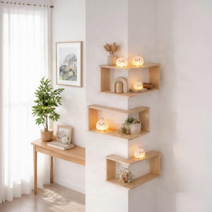 Op de afbeelding: Drie lichtgekleurde houten hoekplanken verticaal gemonteerd aan een witte muur. De planken bevatten kleine, gloeiende, ronde kuikenvormige lampen, een houten boog, een kleine potplant en decoratieve items in een lichte, luchtige kamer.