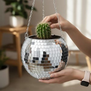 Puede incluir: Una maceta colgante de bola de discoteca con un pequeño cactus. La maceta esférica está cubierta de azulejos espejados y suspendida por una cadena plateada. El cactus es verde con pequeñas espinas.