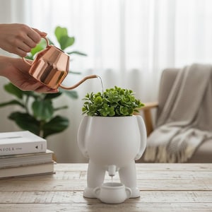 Puede incluir: Una maceta de cerámica blanca con forma de persona, con una maceta pequeña debajo. Contiene una planta verde y se riega con una regadera de cobre. La maceta tiene un diseño único y caprichoso.