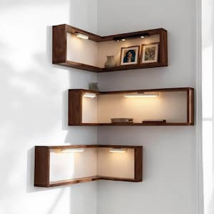 Op de afbeelding: Drie L-vormige hoekplanken van donker hout met witte achterpanelen. Elke plank is voorzien van geïntegreerde LED-verlichtingsstrips die het interieur verlichten. De bovenste plank bevat twee ingelijste foto's en een kleine vaas.