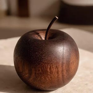 May include: A decorative apple sculpture carved from dark brown wood with a visible natural grain pattern. The object features a smooth, rounded body and a single, thin, curved wooden stem extending from the center of the top indentation.