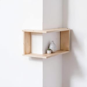 May include: A light-colored wooden corner shelf featuring a two-tier design that wraps around the wall edge. The shelf is constructed from plywood with visible layered edges. A small white ceramic vase and a tiny potted succulent sit on the shelf.