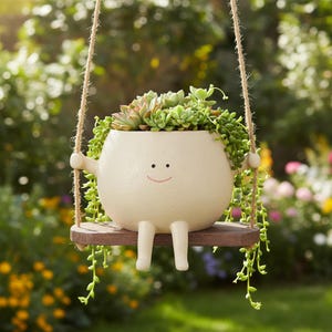 Könnte beinhalten: Ein cremefarbener Pflanztopf in Form eines lächelnden Charakters, gefüllt mit grünen Sukkulenten. Der Topf hat Arme und Beine und hängt an einer Seilschaukel. Der Holzschaukelsitz verleiht dieser einzigartigen Gartendekoration eine rustikale Note.